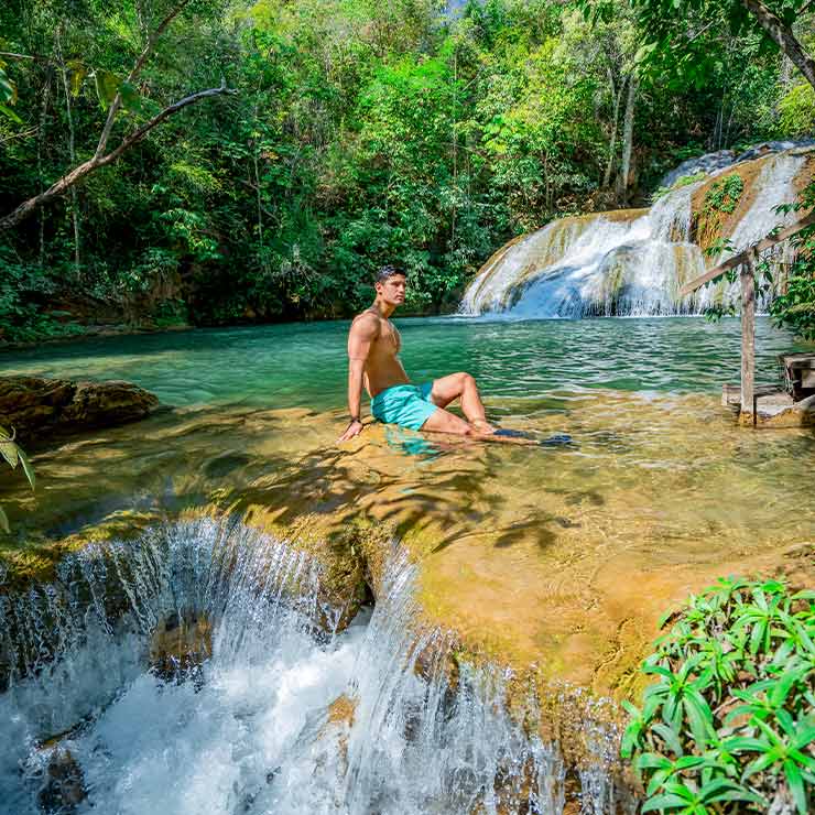 Homem sentado em uma pedra ao lado de uma cachoeira em uma piscina natural, cercado por vegetação densa.