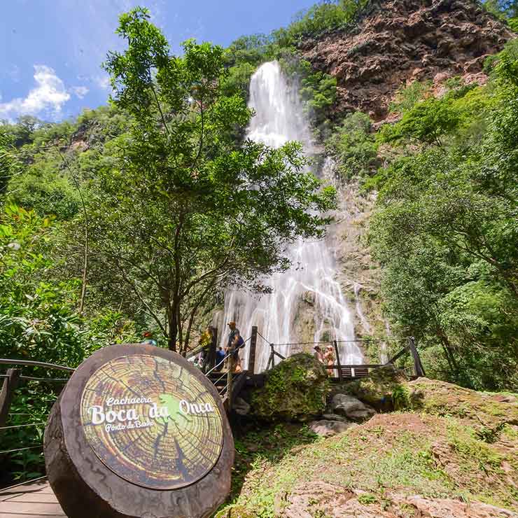 A Cachoeira Boca da Onça desce majestosa entre vegetação exuberante, com placa indicativa em destaque no primeiro plano