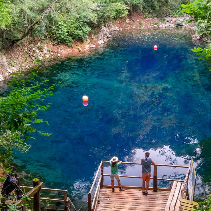 Duas pessoas observam uma lagoa de águas azuis e transparentes, rodeada por vegetação densa e pedras