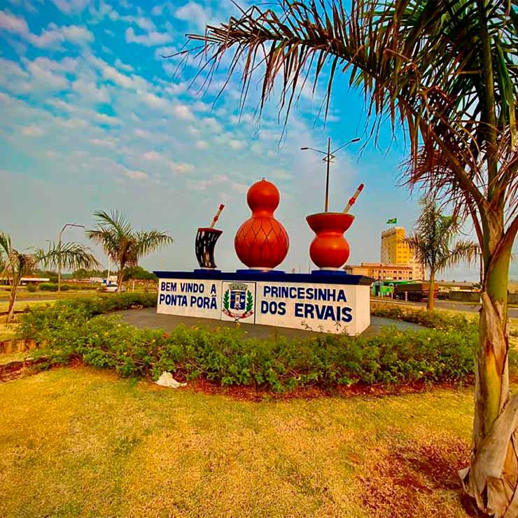 Placa de boas-vindas a Ponta Porã, "Princesinha dos Ervais", com cuias de chimarrão, cercada por palmeiras e plantas