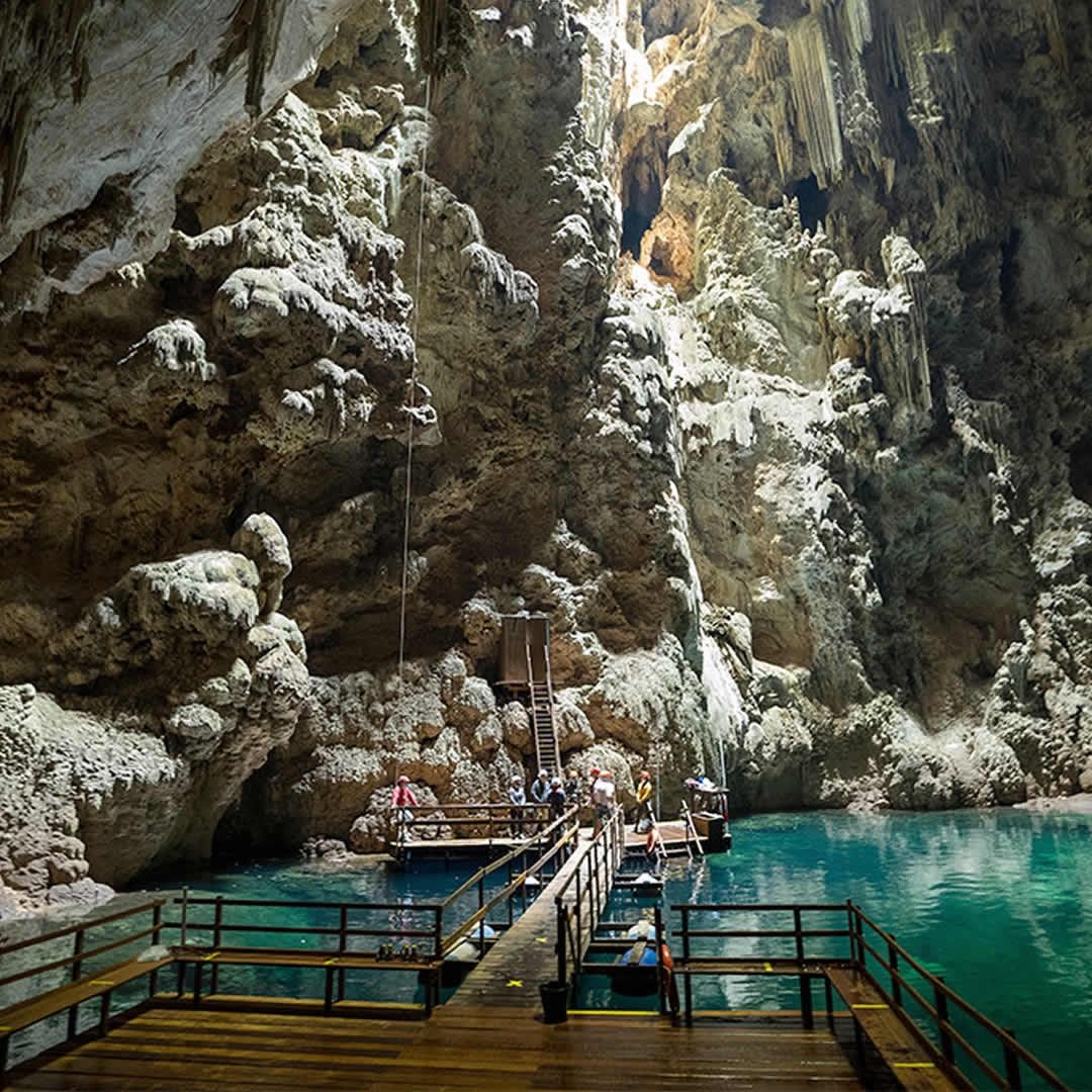 Caverna com águas turquesa, formações rochosas e plataforma de madeira para turistas explorarem a caverna e descerem até a água cristalina.