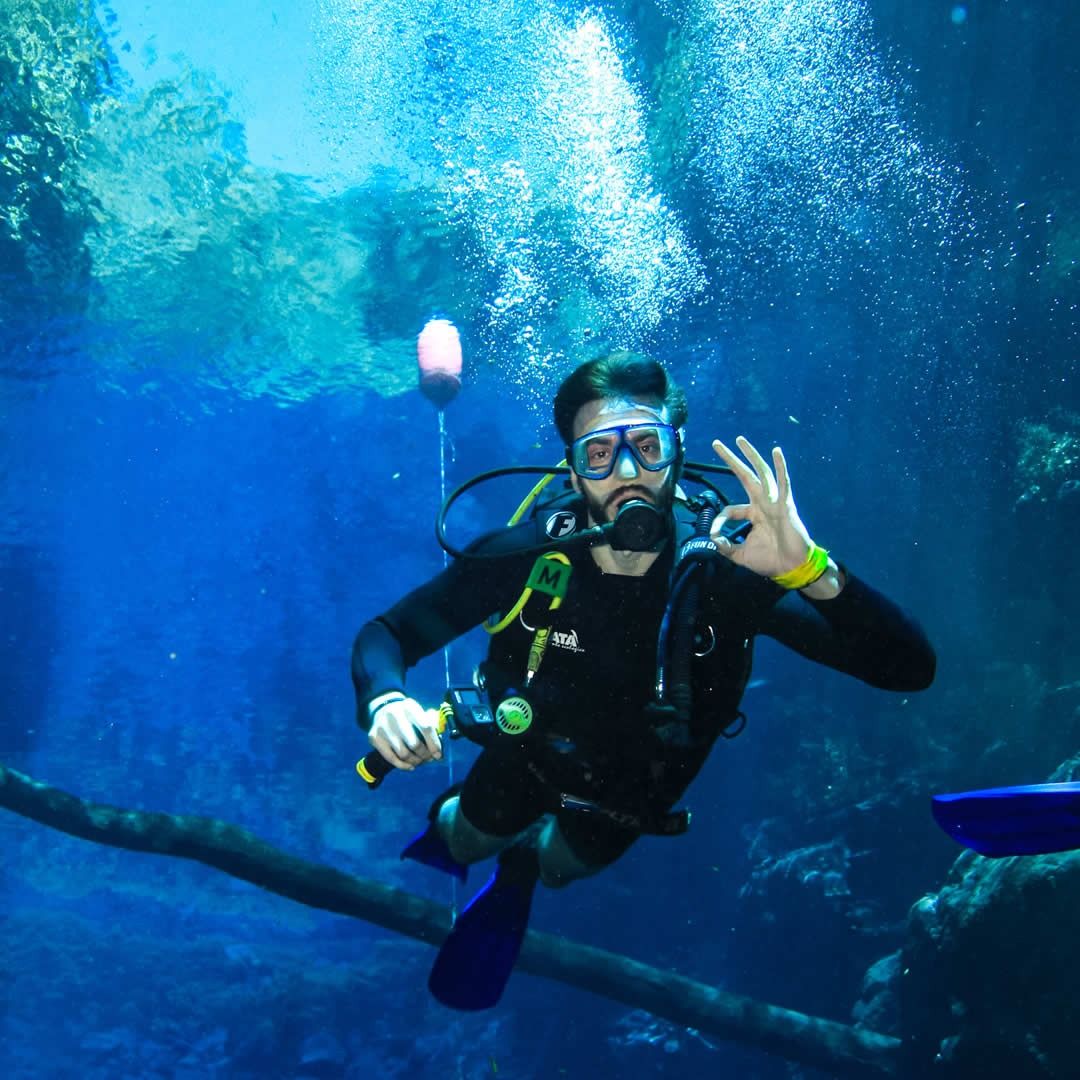 Pessoa fazendo mergulho com equipamento de mergulho em ambiente aquático, fundo azul e bolhas de água ao redor.