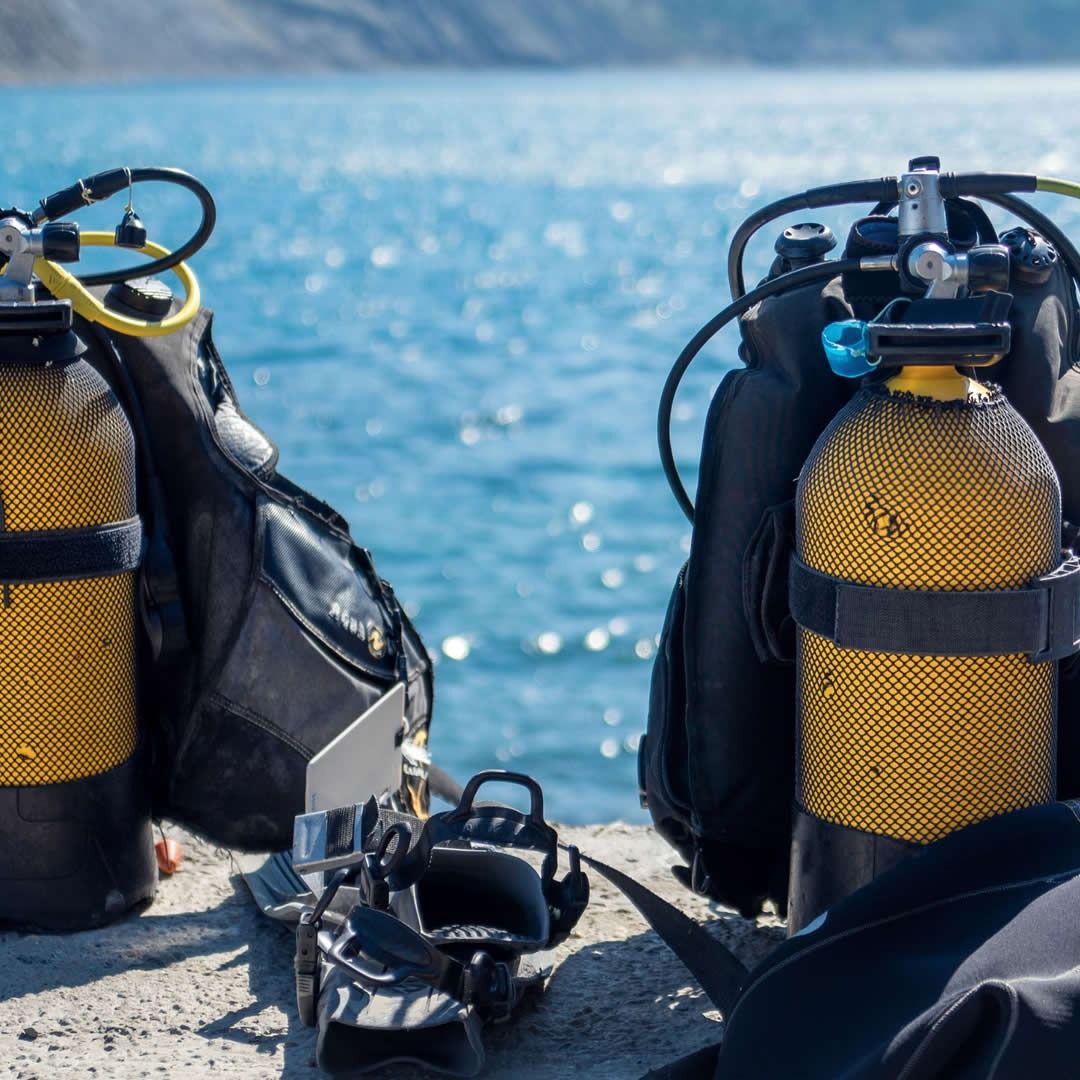 Preparação para mergulho com equipamentos de snorkel na areia, incluindo cilindros de oxigenio, máscara de mergulho, colete de mergulho e bolsas junto ao mar.
