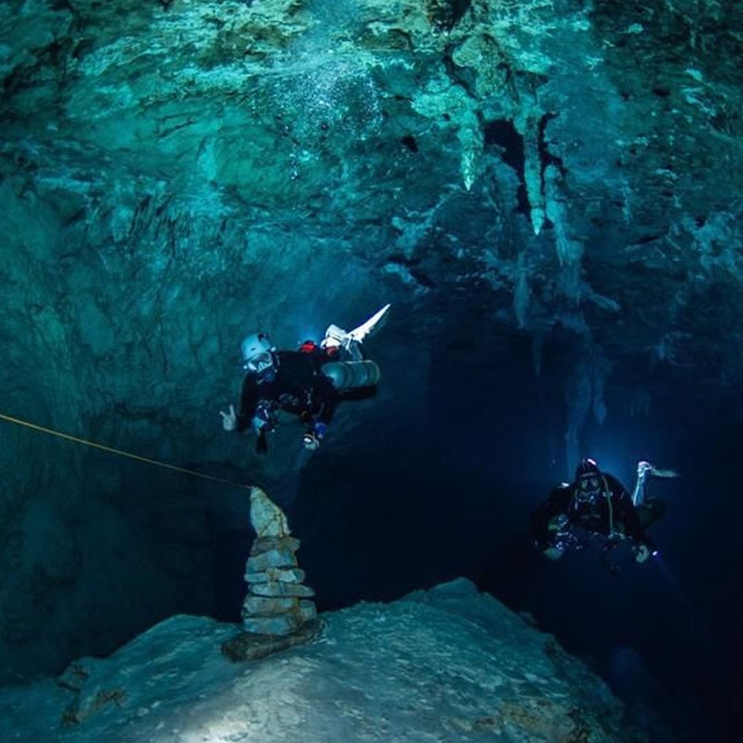 Exploradores de cavernas mergulham no interior de uma caverna profunda de cor turquesa, explorando formações rochosas e câmaras subterrâneas, aventurando-se e praticando espeleologia em um ambiente misterioso.