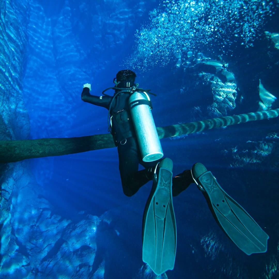 Homem praticando mergulho em um ambiente aquático com equipamento completo, incluindo cilindro de oxigênio e nadadeiras, explorando um cenário subaquático com formações rochosas e água azul clara.