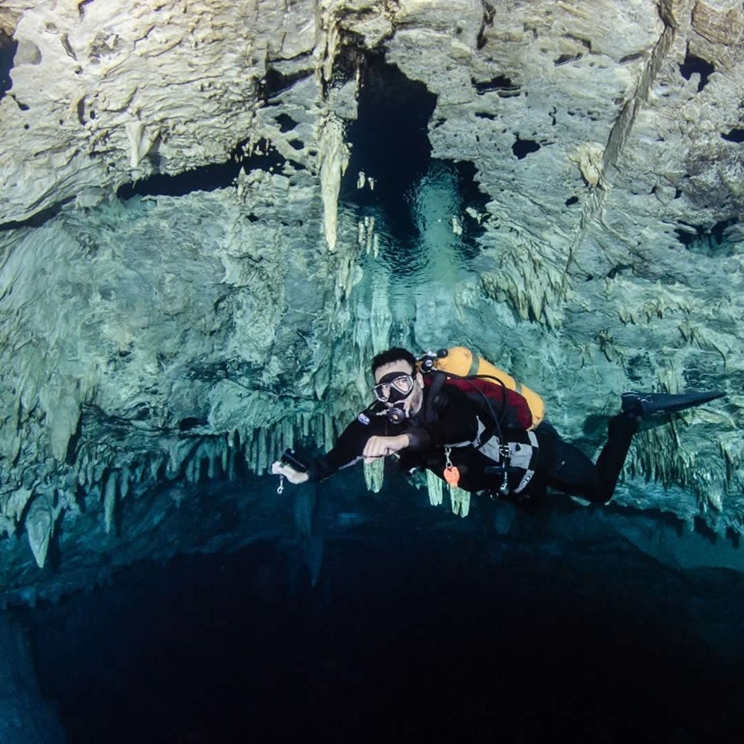 Cave explorer exploring a stunning underground cavern with stalactites and stalagmites, wearing scuba gear and flashlight, capturing the beauty of the cave interior.